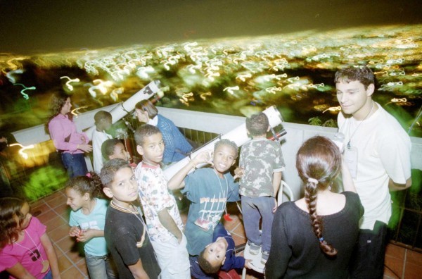 Dezembro/03 - Observatório Astronômico da UFMG leva a ciência do céu ao Aglomerado da Serra - Marcelo Sant'Anna (Estado de Minas)
     
    
    
        Matéria publicada no jornal Estado de Minas em 10/11/03:
        “O que é que tem de mais bonito para ver no céu?”, perguntou o professor. “A Lua!”, responderam as crianças em coro. Assim começou uma aula diferente na Rádio Favela, no Aglomerado da Serra, região sul de Belo Horizonte. . . . O professor usou recursos de multimídia para mostrar às crianças como que é que uma sombra esconde a Lua durante algumas horas no céu. . . .
        O fundador da Rádio Favela, Mizael Santos, acha que a iniciativa de levar a ciência para a comunidade do Aglomerado da Serra está além do conhecimento. “É uma integração entre o asfalto e o morro”, diz. . . .
        Depois da aula do professor do Departamento de Física da UFMG, as crianças foram ver a Lua de perto. “Você podia ver Zé, ela muda de lugar”, disse André Alves de 11, ao amigo, entusiasmado com a imagem. “A Lua é bonita, cheia de detalhes”, disse Kely Janilson, de 13. Claudiane, de oito, ficou encantada com as crateras. “Eu vi um monte de pequenos buracos. A Lua é redonda e amarela”, disse. . . . (repórter Isadora Camargos).
       
        